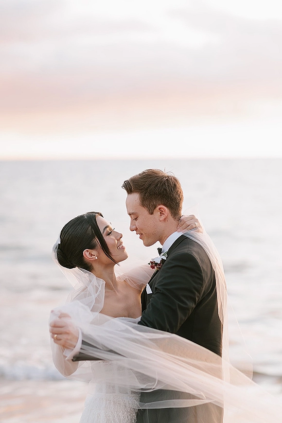 Couple portrait of bride and groom embracing on the beach at sunset, her veil blowing in the ocean breeze near the horizon