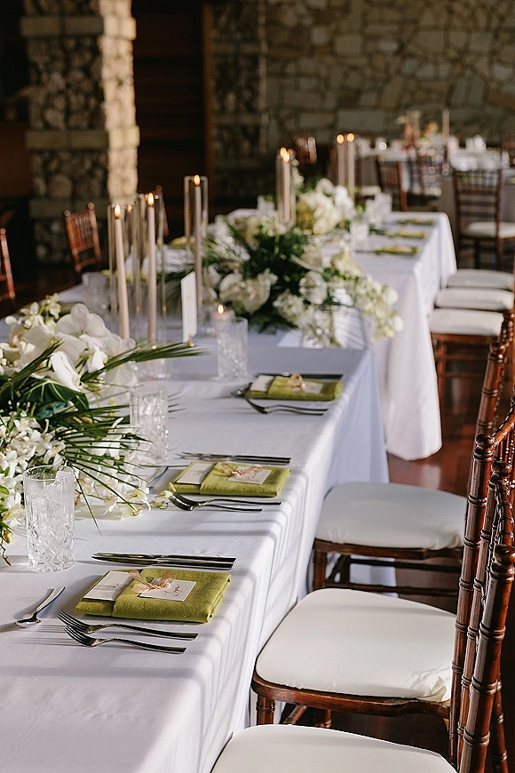 Reception tablescape with head table decor featuring greenery garland, white florals, taper candles, and green napkins against a stone wall indoors