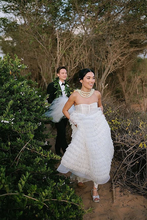 Wedding couple portrait of bride and groom walking on a forest path, her tiered ruffle dress with tulle train and his tux with greenery lei