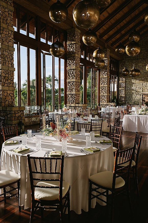 Reception tablescape with round wedding tables, white linens, floral centerpieces in cylinder vases, and amber pendant lights by a stone fireplace