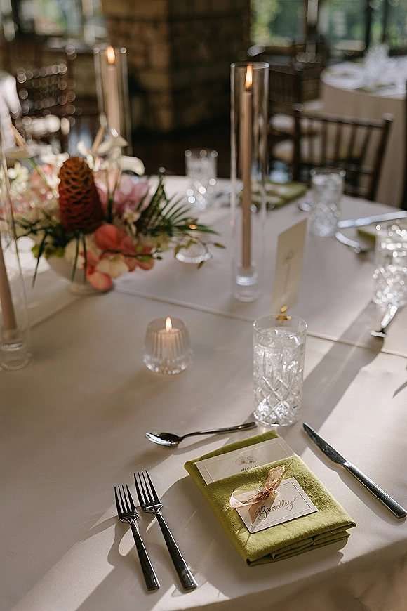 Reception tablescape with wedding place setting featuring a green napkin, ribboned place card, menu, crystal glassware, and tall candle centerpiece