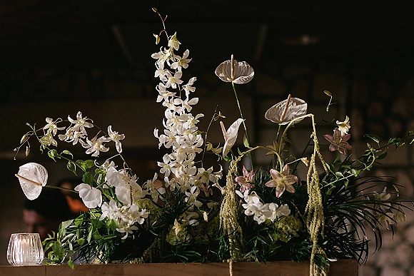 Wedding floral centerpiece with white orchid blooms, anthurium, lilies, trailing amaranthus, and greenery beside a votive candle in a dim venue