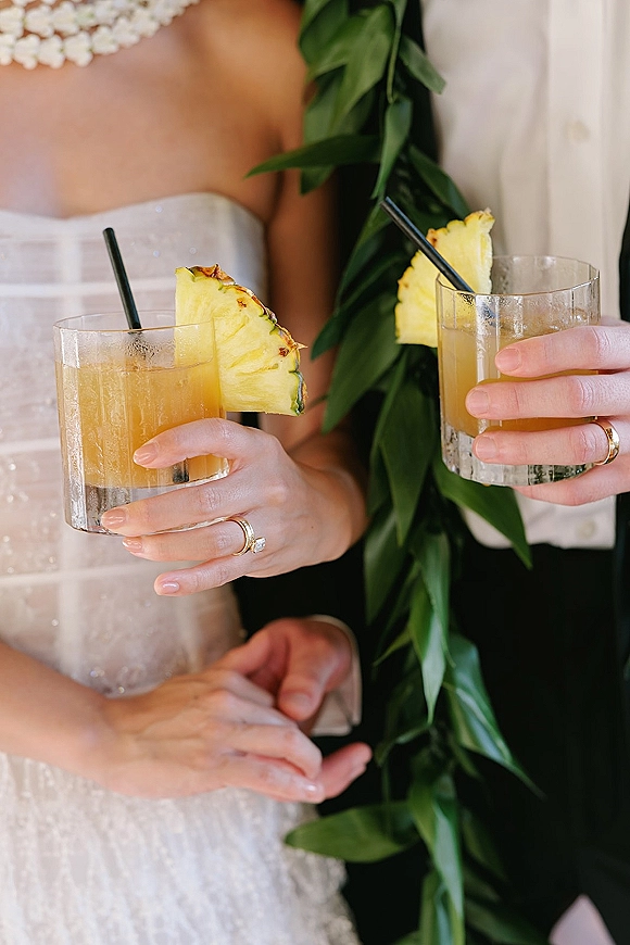 Wedding cocktails held by bride and groom in glass tumblers with pineapple garnish and straws, rings visible against a neutral indoor backdrop