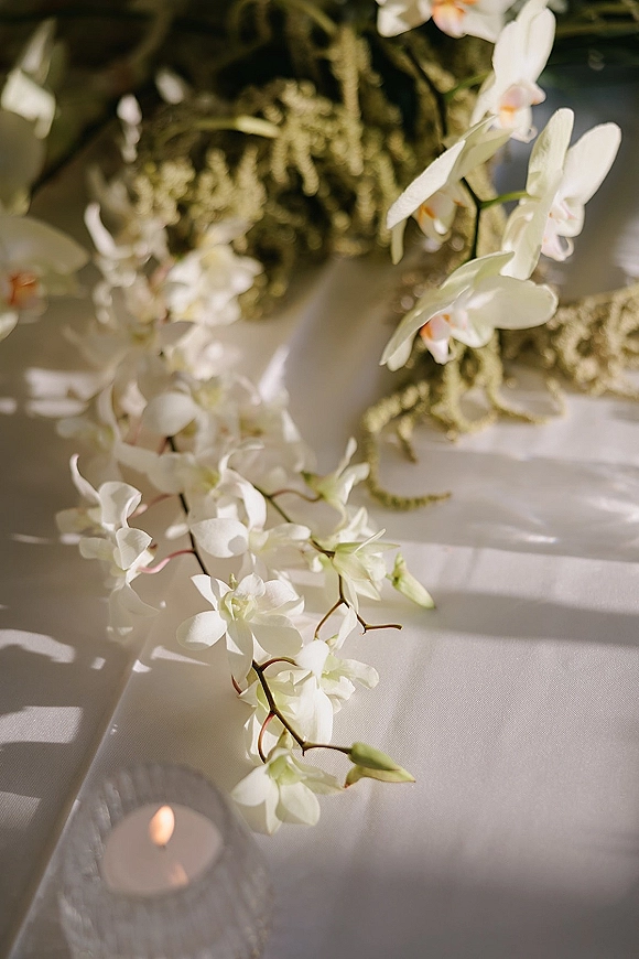 Wedding table decor with white orchids and greenery garland on a white tablecloth, lit by a votive candle with soft shadows