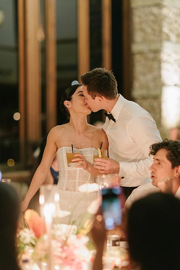 Wedding kiss moment as bride and groom kissing at their reception table, holding cocktails with straws amid candlelight and floral centerpiece