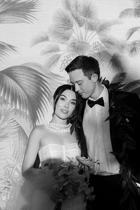 Couple portrait, black and white wedding portrait of bride in strapless gown with floral necklace as groom in tux kisses her forehead by tropical leaf wallpaper