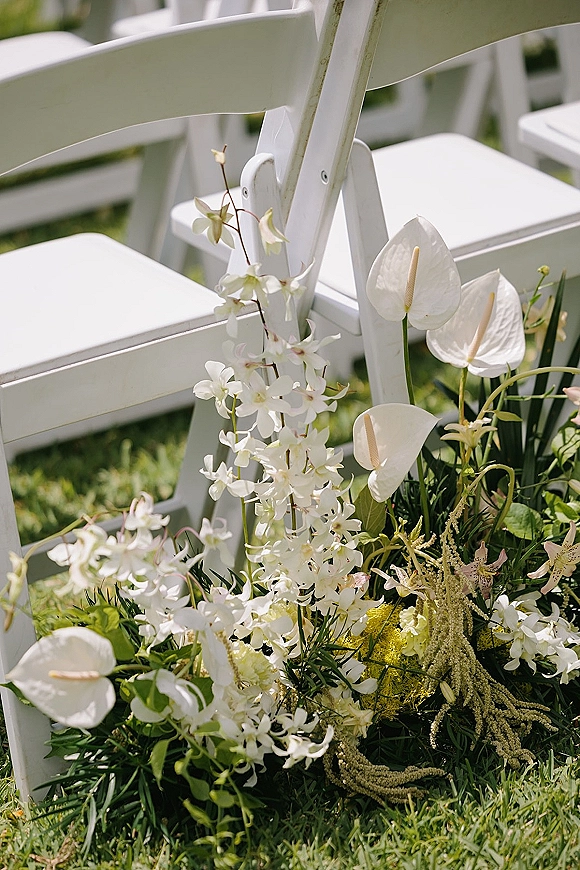 Ceremony aisle flowers and wedding aisle floral arrangement with white orchids and anthurium beside white folding chairs on a grass lawn