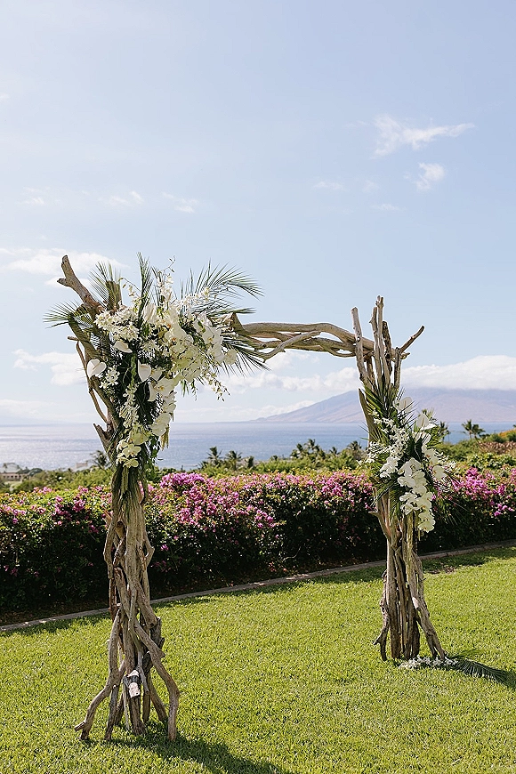 Wedding ceremony arch with driftwood wedding arch design, white orchids and palm fronds on a lawn overlooking the ocean and mountains