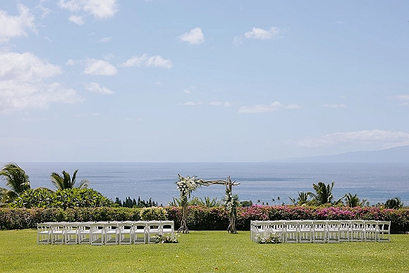 Ceremony setup for an outdoor wedding ceremony with a rustic wood arch and white florals on a lawn overlooking the ocean and palms