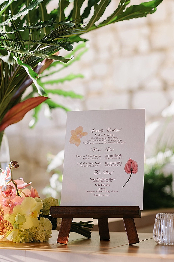 Wedding bar menu on a wooden easel with a drinking glass and tropical greenery, orchids, and hydrangea against a stone wall backdrop