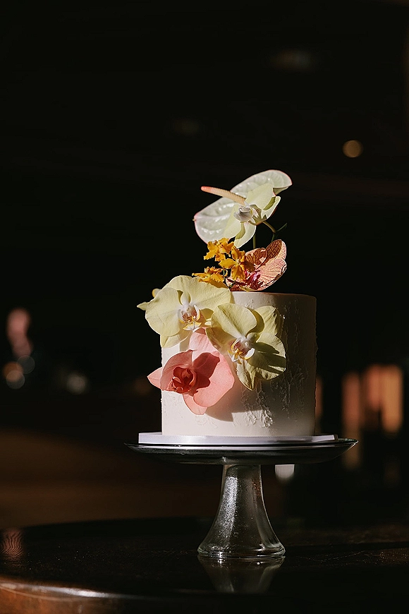 Wedding cake with textured white buttercream topped with orchid and anthurium flowers on a stand against dark reception bokeh lights