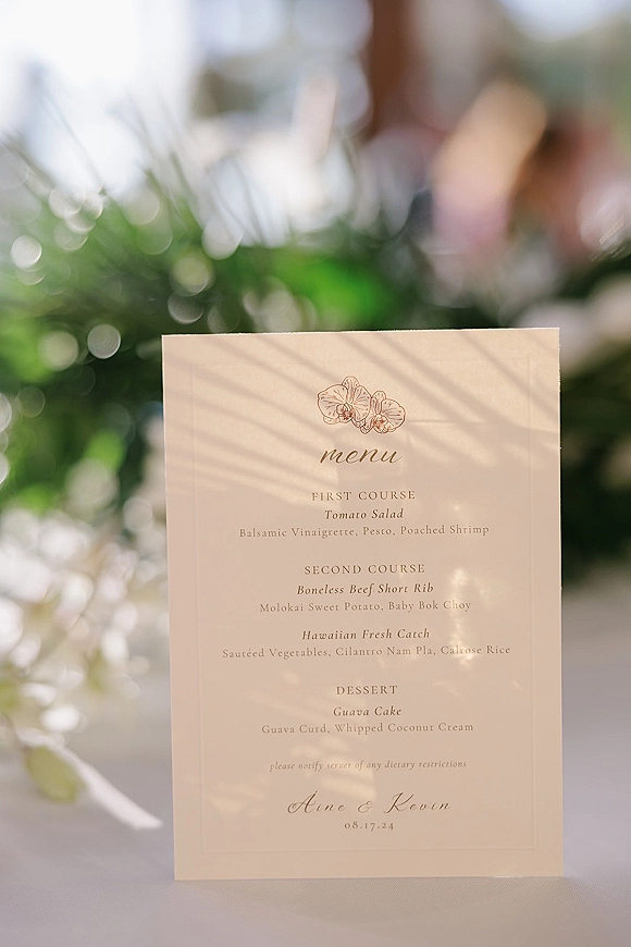 Wedding menu card with orchid illustration and calligraphy typography on a table, set against outdoor greenery with soft bokeh lights