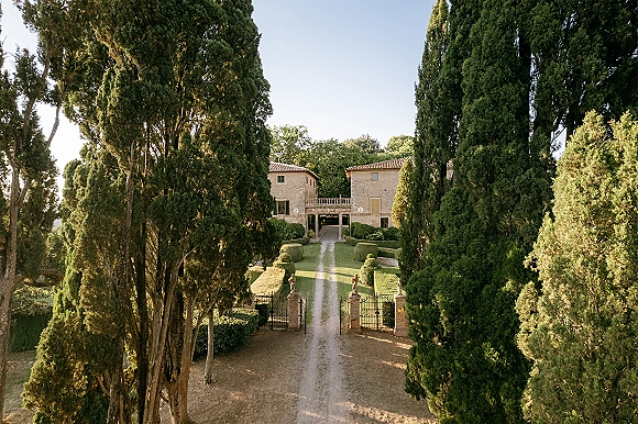 Wedding venue exterior with a stone villa, gravel driveway, and wrought iron gates between brick pillars and manicured hedges
