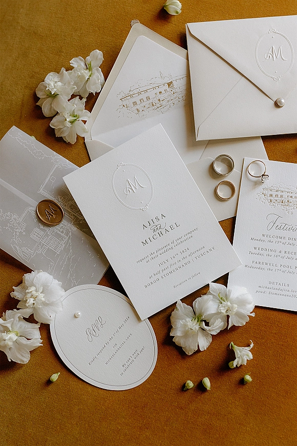 Invitation suite with embossed wedding invitations, wax-sealed envelopes and wedding rings on brown fabric backdrop with a tulip bud