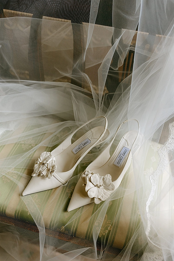 Bridal shoes, ivory wedding heels with 3D flower appliques resting on an upholstered chair, styled with tulle veil and lace dress