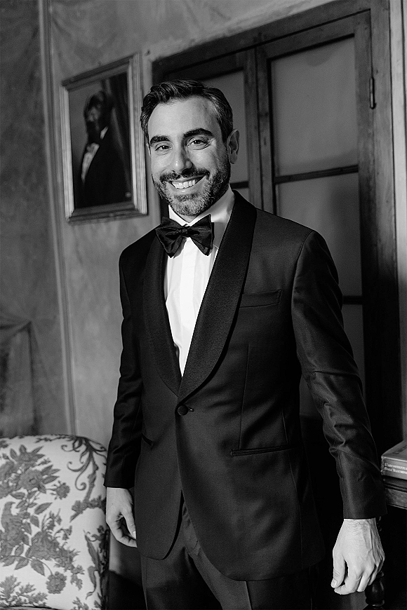 Groom portrait in a black tuxedo, bow tie, and white dress shirt standing in a vintage room with wood doors and a patterned sofa