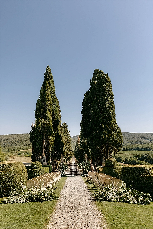 Ceremony aisle design with outdoor wedding aisle lined in white florals and greenery garlands, wood chairs on gravel, garden gate and cypress trees beyond
