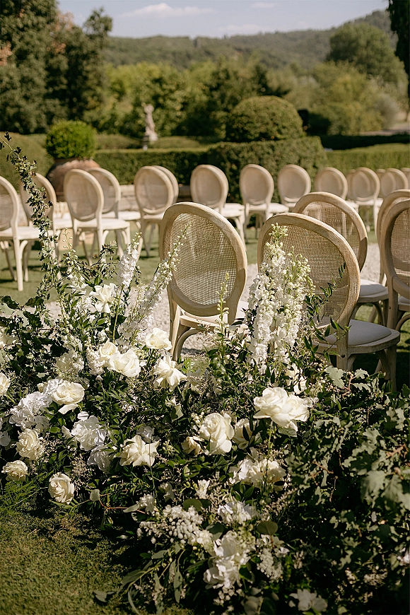 Outdoor ceremony setup with white floral arrangements and greenery garland lining an aisle of cane back chairs on a manicured garden lawn