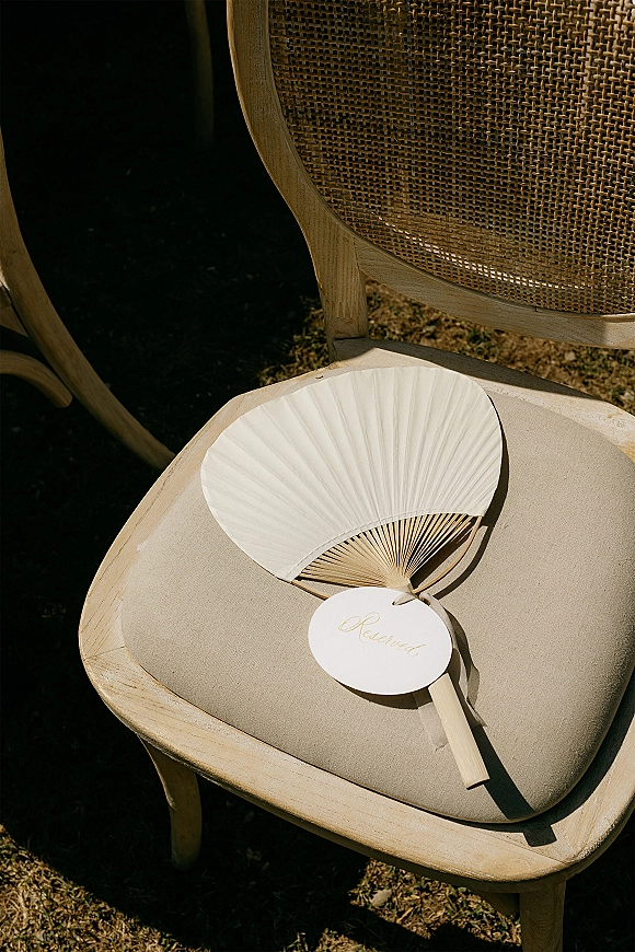 Wedding ceremony fan with a reserved seat sign tag on a wooden handle, resting on a cane-back chair in sunlit outdoor grass