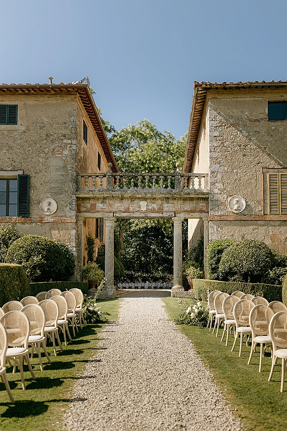 Ceremony setup with outdoor ceremony aisle lined in greenery, symmetrical chairs, and floral markers in a stone courtyard villa under blue sky