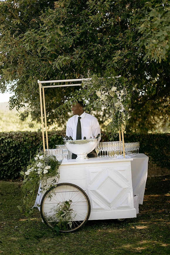 Wedding bar setup on a white bar cart with champagne coupe glasses, wine bottles and greenery, set on a lawn under a large tree canopy