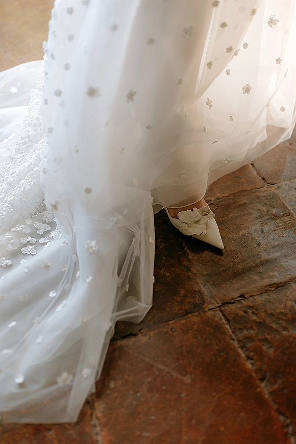 Bridal shoes, ivory wedding heels with pointed toes and floral appliqué beside a sheer embroidered tulle gown hem on a stone floor