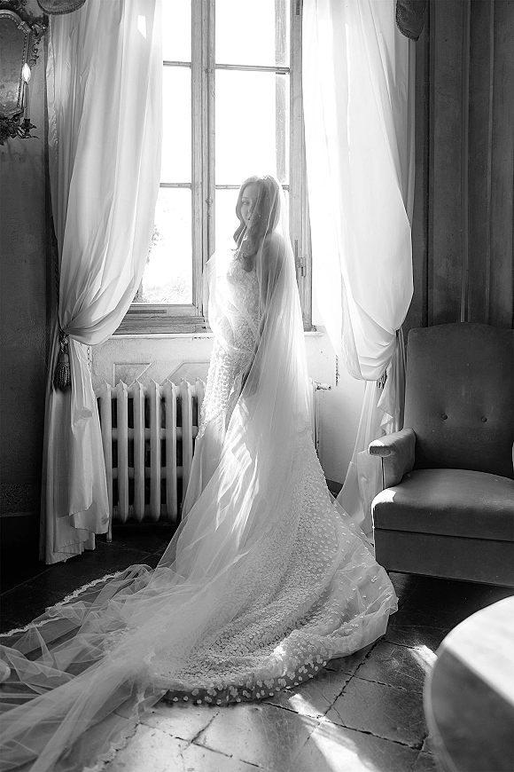 Bridal portrait of a bride in veil covering her face, wearing a lace wedding dress with long train by a large window with sheer curtains