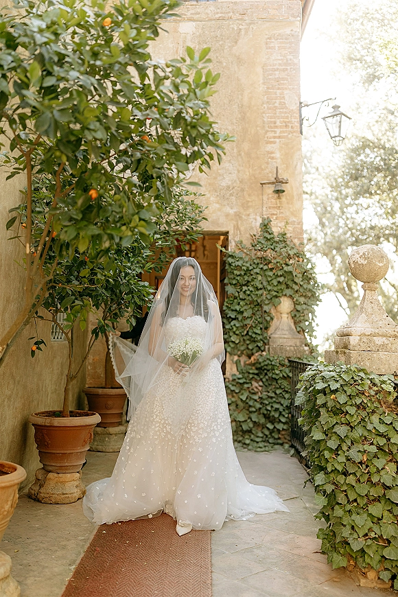 Bridal portrait of a bride with veil draped over her face, holding a baby’s breath bouquet in a stone courtyard with terracotta pots.