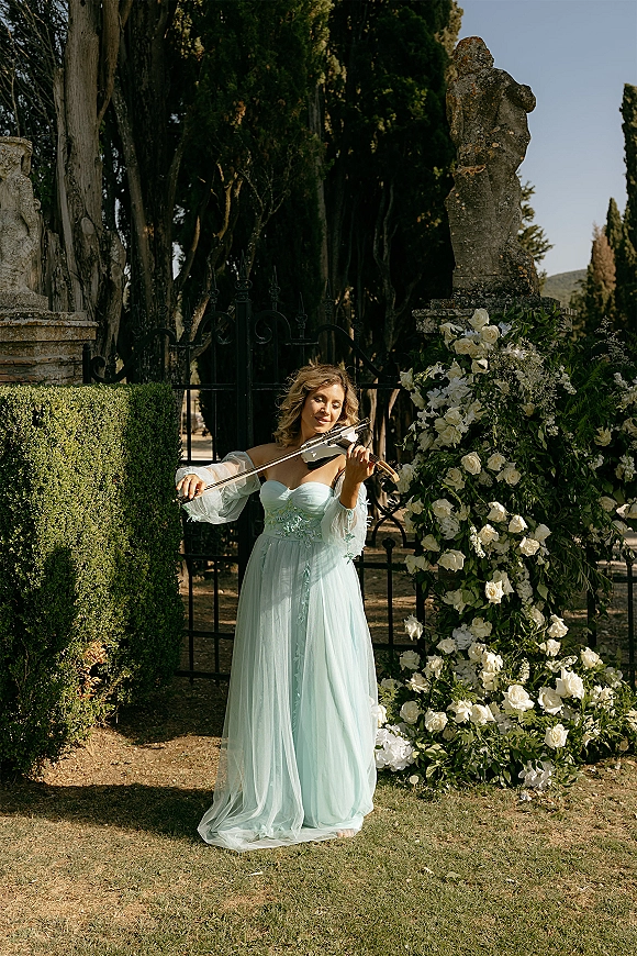 Wedding musician playing violin in a light blue off-shoulder dress beside a white rose floral arch and wrought iron gate on a garden lawn