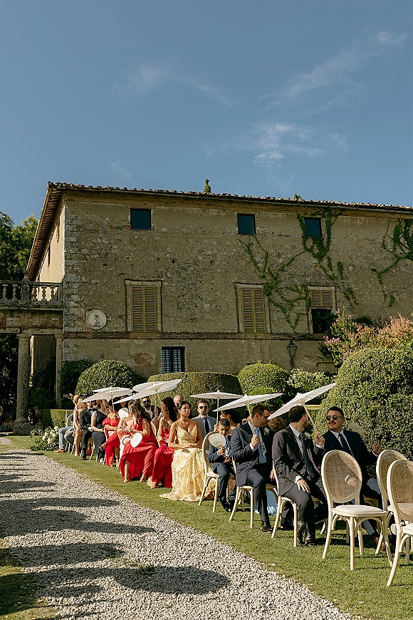 Wedding guests seated at an outdoor ceremony, shaded by white parasols, watching beside a gravel aisle near an ivy-covered stone villa