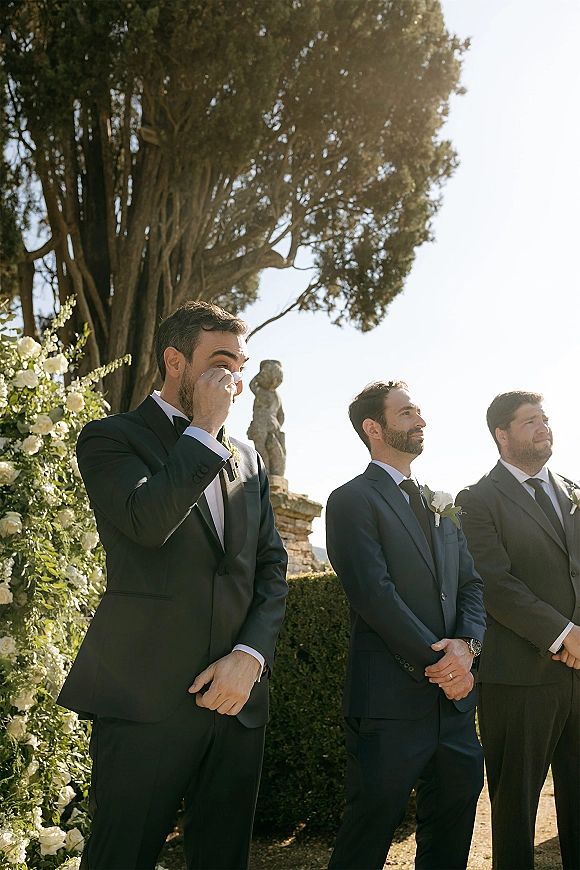 Groom emotional moment as he wipes tears in a black tuxedo with white rose boutonniere, standing in a bright outdoor garden ceremony setting