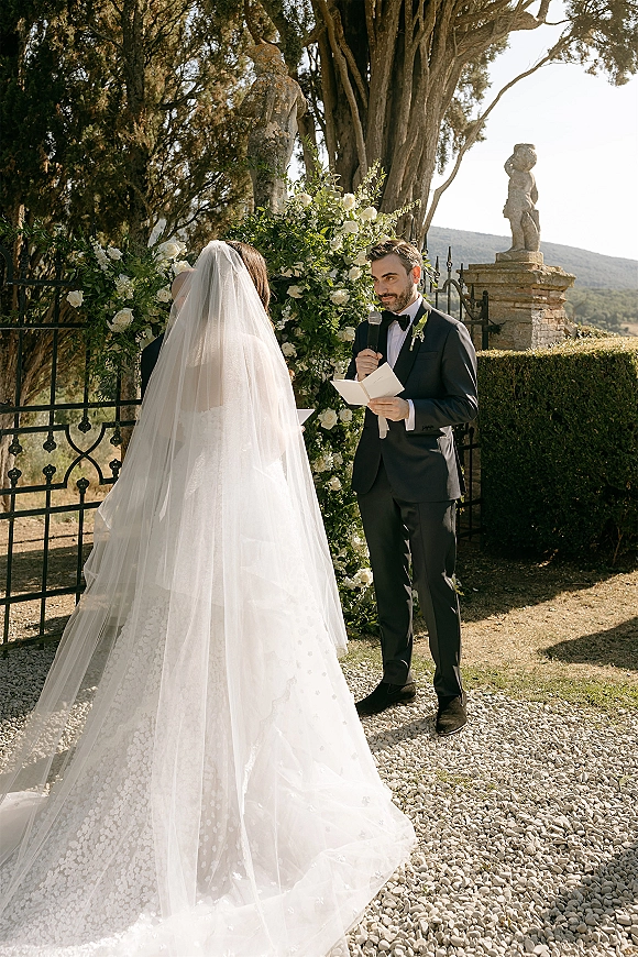 Wedding vows as the groom reading vows into a microphone beside the bride’s veil, under a white rose arch by a wrought iron gate