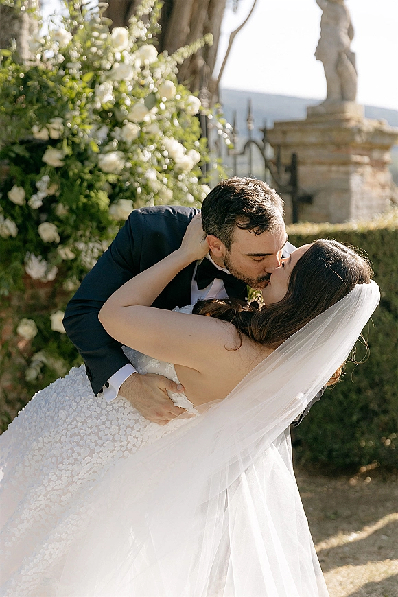 Wedding kiss portrait of groom dipping bride in lace dress, long veil flowing, against a floral wall with sunlit garden hedges