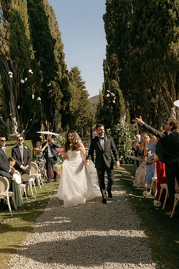 Wedding recessional as bride and groom walking aisle through a tree-lined garden, flower petals falling, veil flowing and guests cheering under blue sky