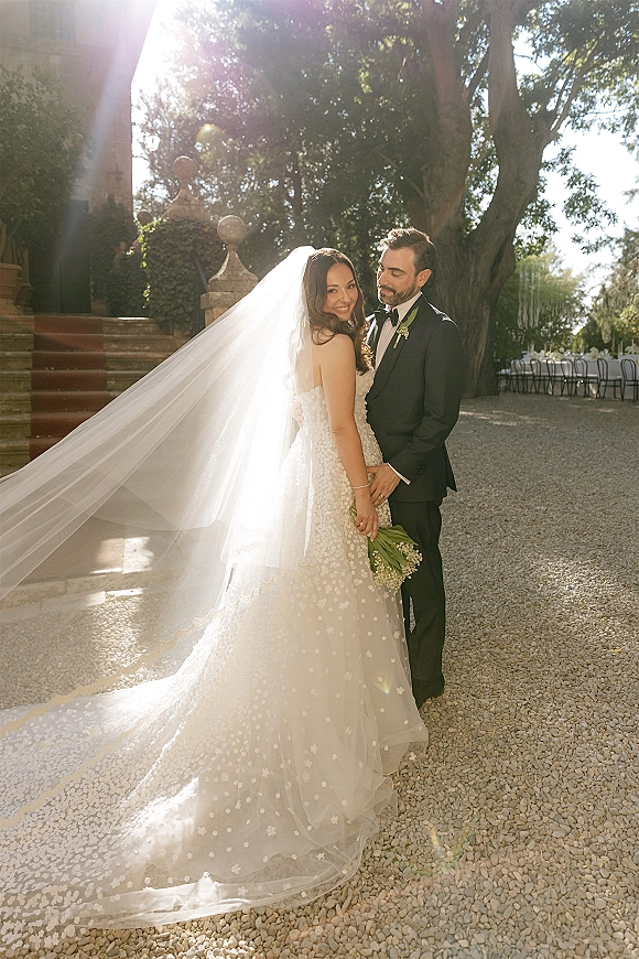 Couple portrait of bride and groom embrace on stone steps, her long veil trailing as she holds a lily of the valley bouquet in sun flare