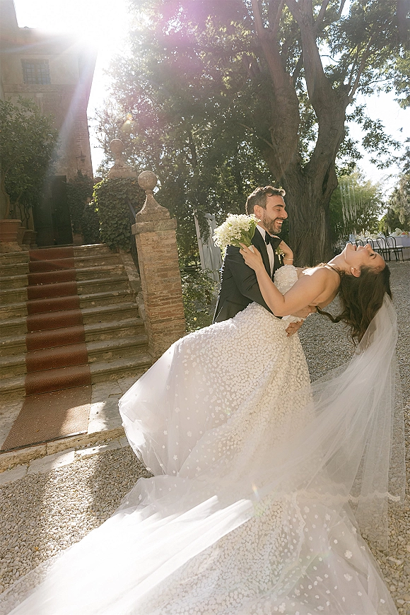 Wedding couple portrait of groom dipping the bride as she laughs with bouquet and veil, by ivy and brick pillars in sunlit courtyard