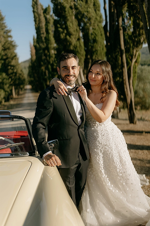 Couple portrait of bride in a sparkly wedding dress adjusting groom’s bow tie beside a vintage convertible on a cypress-lined road