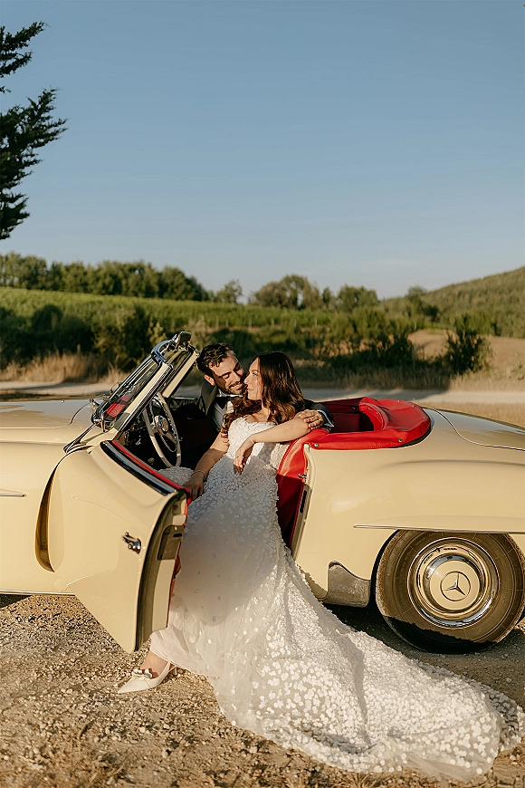 Couple portrait beside a vintage car wedding photo, bride in lace dress and groom in tuxedo by open convertible on vineyard road