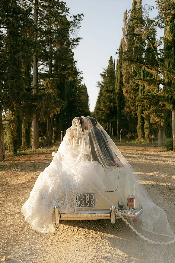 Wedding couple portrait of bride and groom in car kissing under a wedding veil on a vintage convertible along a tree-lined dirt road