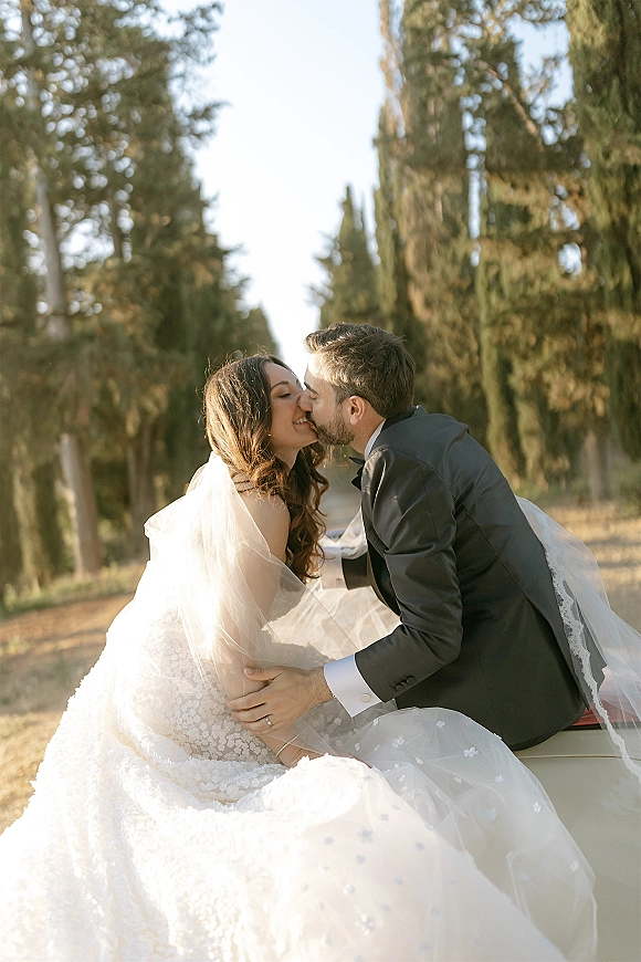 Wedding kiss portrait of bride and groom kissing on a car hood, cathedral veil blowing over lace dress on a sunlit tree-lined road