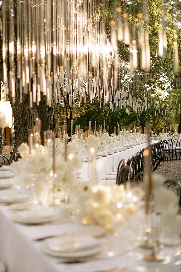 Reception tablescape with a long banquet table setup, white florals and taper candles in glass holders under tube lights in a garden setting