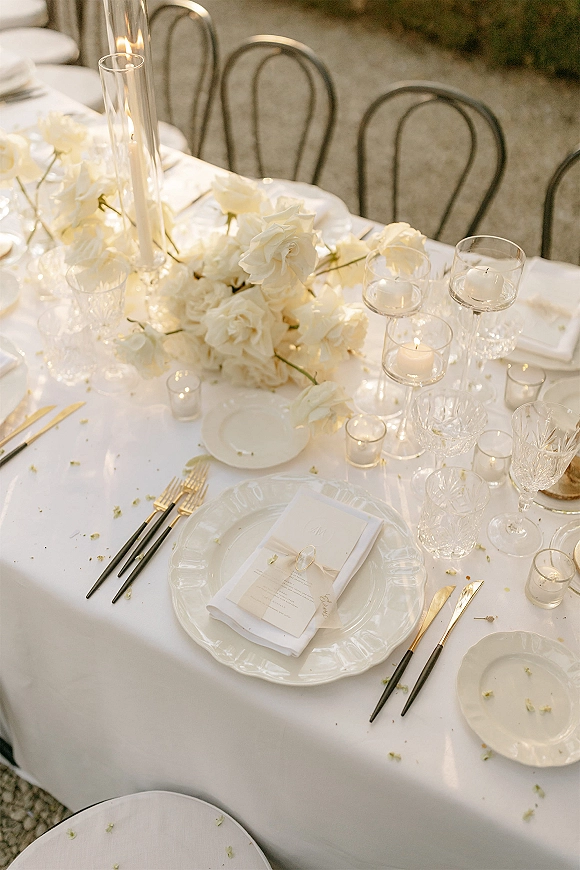 Reception tablescape with a white wedding tablescape featuring white rose centerpiece, taper candles in glass cylinders, and black and gold flatware outdoors