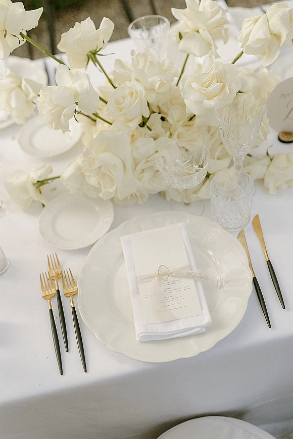 Reception tablescape with white rose centerpiece, gold flatware, crystal goblets, and menu on napkin on an outdoor stone backdrop