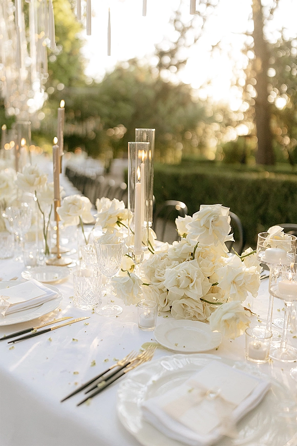 Reception tablescape with white wedding tablescape styling, white roses and taper candles in glass holders beneath hanging pendants outdoors at sunset