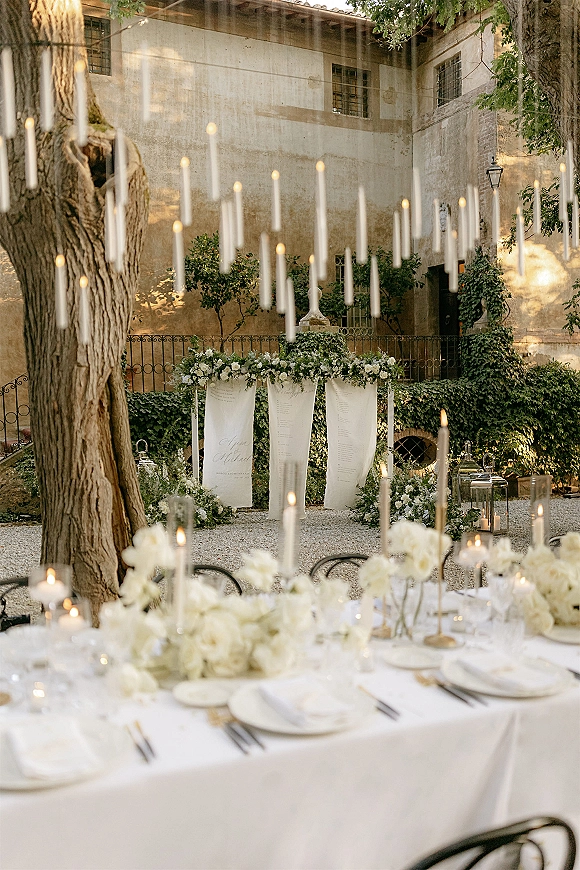 Wedding reception decor with outdoor reception decor, long white banquet table under hanging taper candles in an ivy-walled stone courtyard with lanterns