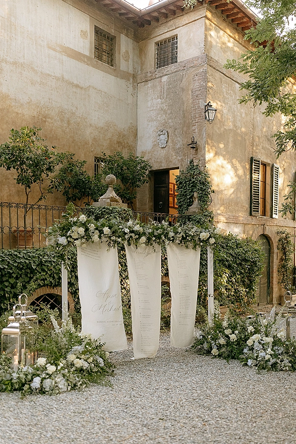 Wedding seating chart on hanging fabric panels with greenery garlands and white florals, set on a villa courtyard railing with lanterns