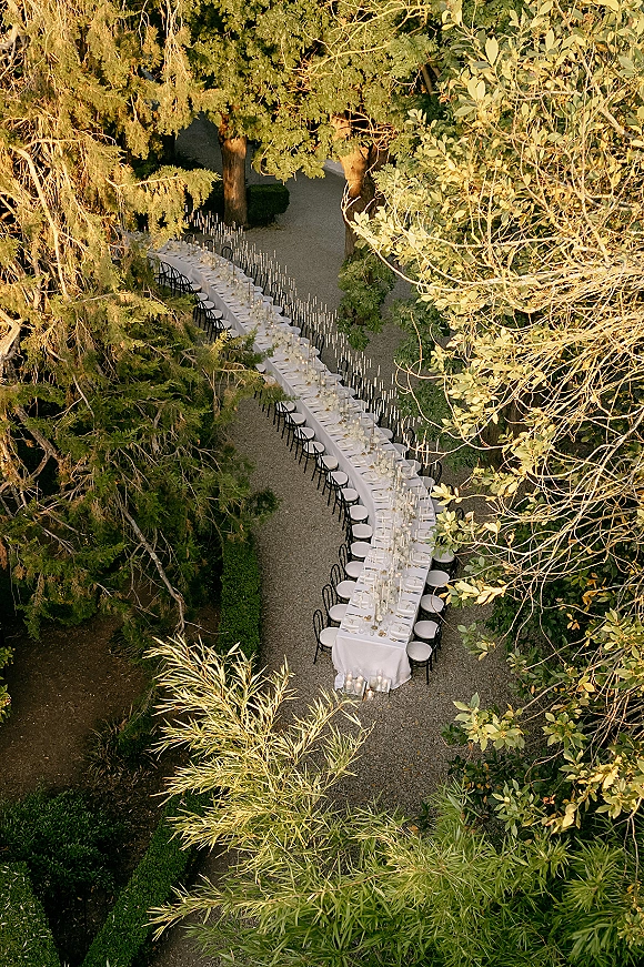 Reception tablescape with outdoor banquet table set in white linen, black chairs, place settings and taper candles along a gravel garden path