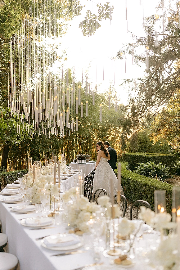 Reception tablescape with an outdoor wedding tablescape of white florals, taper candles, and black bentwood chairs in a sunlit garden setting
