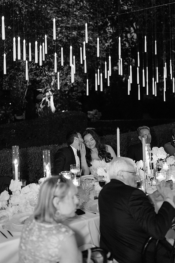 Reception moment of bride and groom at head table whispering under hanging taper candles, surrounded by floral centerpieces in a night garden setting