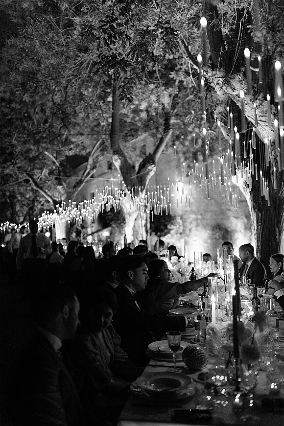 Reception dinner at a long banquet table wedding with taper candles, floral centerpieces and string lights under trees in a garden at night